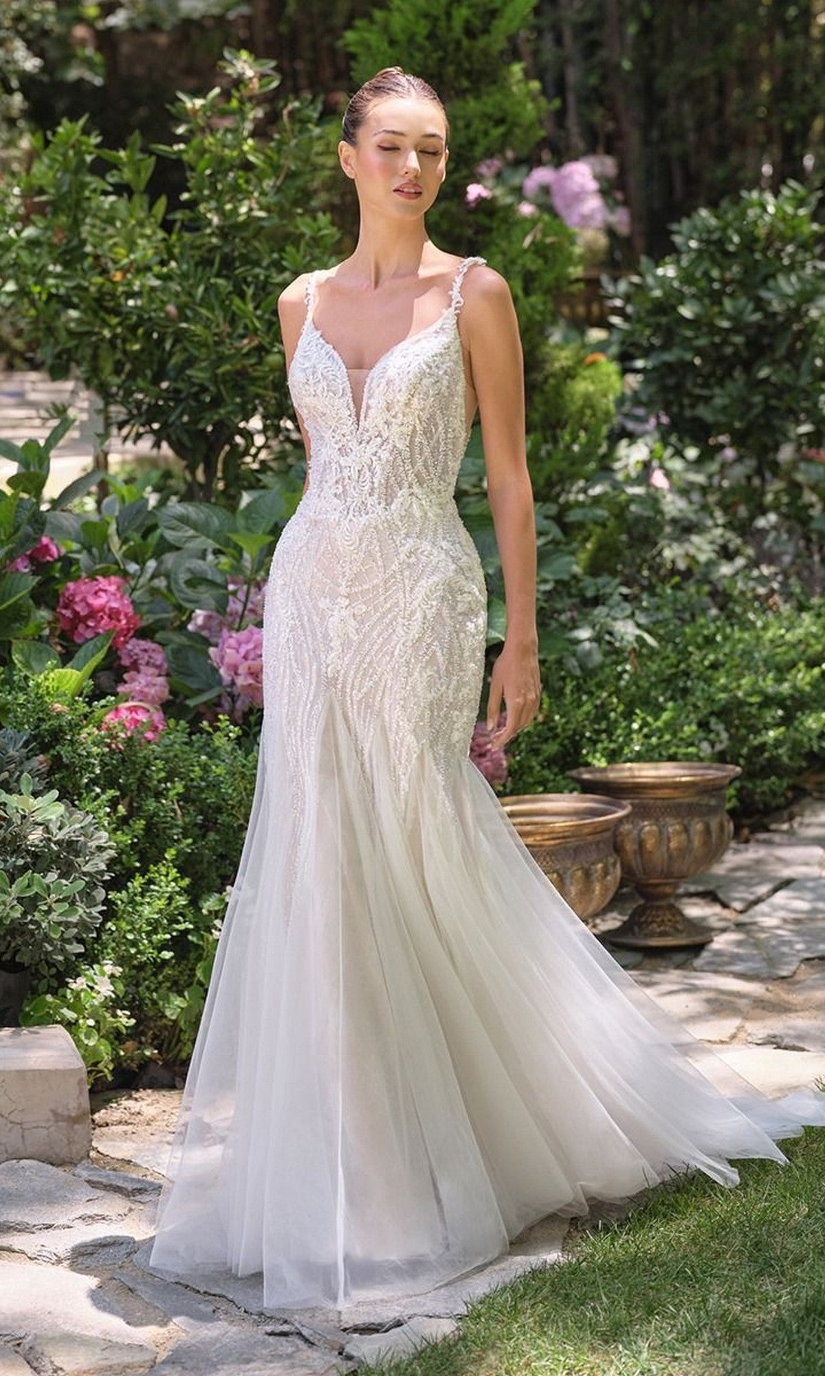 A woman stands in a garden wearing the Andrea and Leo WL066 long wedding dress, a fitted, sleeveless white fit-and-flare gown featuring a deep neckline and lace detailing.